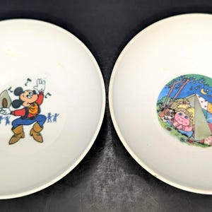 May include: Two round, off-white bowls. The left bowl features a graphic of Mickey Mouse wearing a red shirt, blue pants, and brown boots, holding a hat. The right bowl features a colorful illustration of a doll camping in a tent at night.