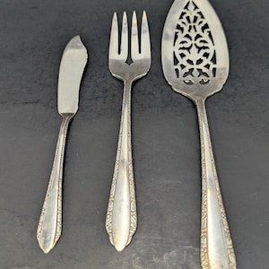 May include: Three pieces of vintage silverware are displayed against a dark background. The set includes a butter knife, a fork, and a serving spoon. The silverware has ornate detailing along the handles and a decorative pattern on the serving spoon.