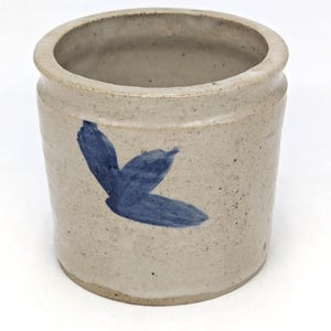 May include: A cylindrical stoneware crock featuring a speckled light beige finish and a hand-painted blue leaf motif on the exterior. The vessel has a thick, rounded rim and a wide opening, showcasing a simple, functional design with visible clay texture.