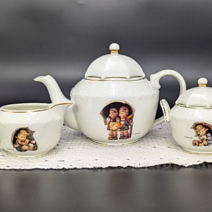 May include: A white porcelain tea set with a delicate floral design. The teapot, creamer, and sugar bowl feature charming illustrations of children under an umbrella.