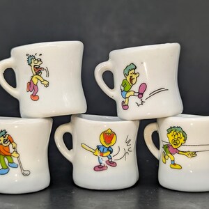 May include: Five small white ceramic mugs with cartoon figures in various poses. Each mug has a handle and a unique illustration in bright colors. The figures are engaged in activities like golfing, running, and batting.
