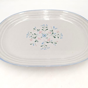 May include: An oval ceramic serving platter with a light gray finish and a blue rim. The center features a painted floral motif with small pink and blue petals surrounded by green leaves arranged in a symmetrical, star-like pattern.