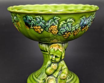 May include: A green ceramic pedestal bowl with a cherub figure on the base. The bowl is decorated with a grapevine design and has a scalloped rim.