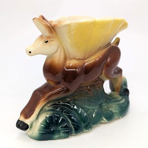 May include: A ceramic deer figurine with a yellow, cream, and brown color scheme. The deer is posed leaping over a green and blue leaf-like base. The figurine has a glossy finish and is likely a decorative item.