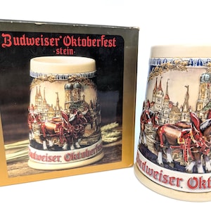 May include: A ceramic beer stein featuring a relief illustration of a horse-drawn wagon in front of a city skyline, with the text Budweiser Oktoberfest printed in red at the base. The stein is accompanied by its original cardboard display box.