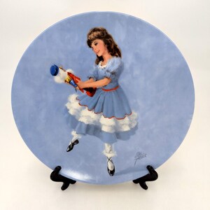 May include: A decorative plate featuring a young girl in a blue dress holding a toy. The plate has a light blue background with a painted image of the girl and toy. The plate is displayed on a black stand.