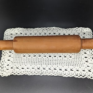 May include: A wooden rolling pin on a white crocheted doily. The rolling pin is brown and has a smooth surface.