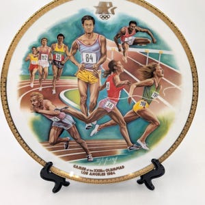 May include: A decorative plate featuring a colorful illustration of Olympic athletes competing in track and field events. The plate has a gold rim and the text "GAMES of the XXIII OLYMPIAD LOS ANGELES 1984".