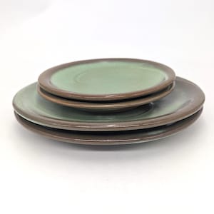 May include: A stack of three round ceramic plates featuring a smooth, sage green center glaze that transitions into a dark brown rim. The plates are layered with the smallest on top and the largest on the bottom, showcasing a consistent color palette.