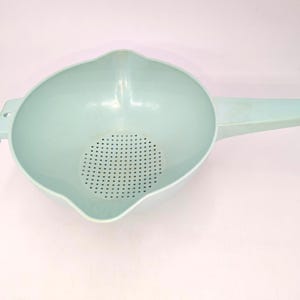 May include: A light blue plastic colander with a handle and a perforated base. The colander has a wide bowl shape with a pouring spout and a handle with a hole for hanging. The kitchen tool is designed for draining liquids from food.
