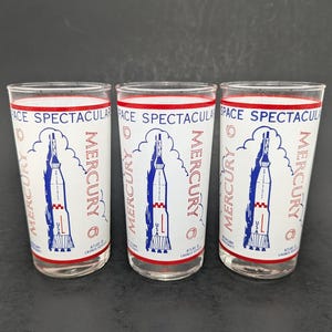 May include: Three vintage drinking glasses with a white background and a red and blue rocket ship design. The glasses have the words "MERCURY" and "SPACE SPECTACULAR" printed on them. The glasses are clear and cylindrical.