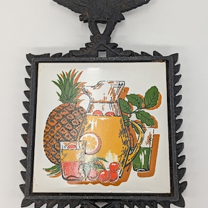 May include: A black cast iron trivet with a decorative eagle at the top. The trivet features a square tile with a colorful illustration of a pineapple, a pitcher of juice, and cherries. The trivet is designed to protect surfaces from hot items.