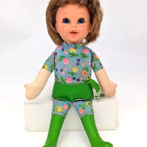 May include: A doll with brown hair styled with a pink bow, featuring large blue eyes and an open mouth. The doll wears a light blue short-sleeved shirt and matching pants with a floral pattern, paired with a green waistband and tall green boots.