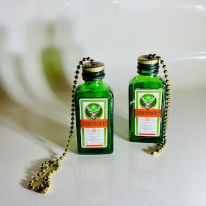 May include: Two miniature green glass bottles with gold chains attached. The bottles are labeled "Jagermeister 56" and feature a stag logo. The bottles are on a white surface.