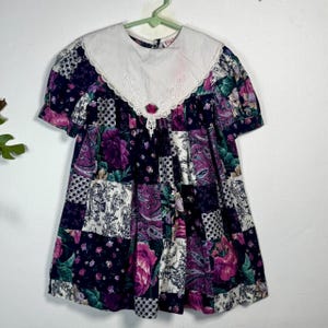 May include: A vintage patchwork dress with a white lace-trimmed collar. The dress features a floral pattern in shades of purple, pink, and green on a navy blue background. The short-sleeved dress is hanging on a green hanger.