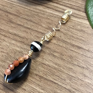 May include: A gold-colored metal hair accessory with a spiral design. It features a black teardrop-shaped bead, a black and white striped bead, and small orange beads. The accessory is designed for hair styling.