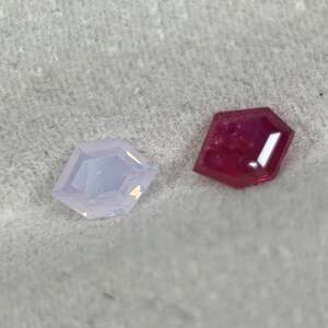 Puede incluir: Dos piedras preciosas facetadas con forma de escudo pentagonal. Una piedra es de un color azul lavanda pálido translúcido, mientras que la otra es de un color rojo rubí profundo y vibrante con facetas internas visibles que reflejan la luz.
