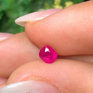Puede incluir: Una pequeña piedra preciosa de talla cojín con un color rojo rosado intenso y vibrante. La piedra presenta múltiples facetas triangulares y trapezoidales en su superficie, creando un aspecto reflectante y brillante.