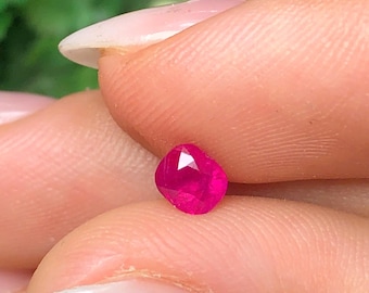 May include: A small, cushion-cut gemstone with a vibrant, deep pinkish-red color. The stone features multiple triangular and trapezoidal facets across its surface, creating a reflective, sparkling appearance when held between two fingers.