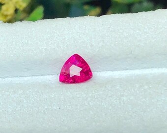 May include: A vibrant, hot pink gemstone cut into a triangular shape with multiple facets that reflect light. The stone features a brilliant, saturated color and a clear, polished surface, showcasing a sharp, geometric silhouette and precise edges.
