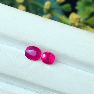 May include: Two loose, faceted rubies with a vibrant, deep pinkish-red colour. The larger stone is oval-shaped, while the smaller stone is cushion-shaped. Both gemstones exhibit clear, reflective surfaces and are positioned on a white surface.