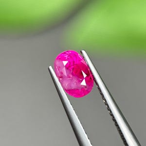 Puede incluir: Una gema de color rosa intenso con corte ovalado sujeta por unas pinzas de metal plateado. La gema presenta una superficie facetada que refleja la luz, mostrando un tono magenta profundo y saturado con inclusiones internas.