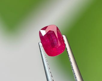 May include: A vibrant, cushion-cut ruby gemstone with a deep red hue and multifaceted surface, held securely by a pair of silver-toned metal tweezers. The gemstone displays internal clarity and light reflection across its polished, geometric facets.