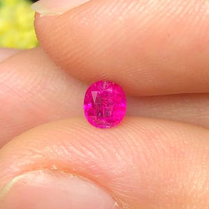 May include: A small, oval-cut gemstone with a vibrant, saturated hot pink colour. The stone features multiple triangular and trapezoidal facets across its surface, creating a reflective, sparkling appearance when viewed under bright lighting conditions.