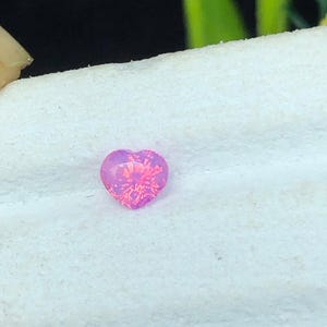 Puede incluir: Una gema rosa en forma de corazón con un interior brillante y texturizado. La gema está colocada sobre un fondo blanco y texturizado. El color de la gema pasa de un rosa intenso a un tono más claro, casi iridiscente.