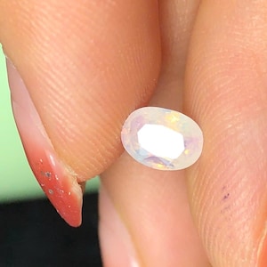 以下が含まれることがあります： 白く虹色の輝きを持つ、楕円形のイリデッセント宝石。 宝石は2本の指の間に挟まれ、研磨された表面と光の屈折を示しています。 貴重な石の美しい例です。