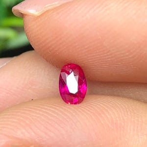 Puede incluir: Una pequeña piedra preciosa de corte ovalado con un color rojo rosado intenso y vibrante. La piedra presenta múltiples facetas triangulares en su superficie que reflejan la luz, creando una apariencia brillante y centelleante.