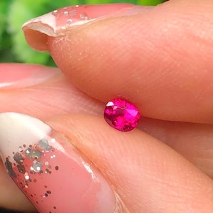 May include: A small, oval-shaped, vibrant pink gemstone. The gem is held between fingers, showcasing its polished surface and intense colour. The background is blurred, highlighting the gem's brilliance.
