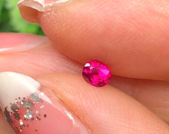 May include: A small, oval-shaped, vibrant pink gemstone. The gem is held between fingers, showcasing its polished surface and intense color. The background is blurred, highlighting the gem's brilliance.