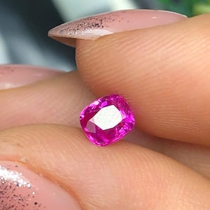 May include: A vibrant, cushion-cut pink gemstone with a faceted surface that reflects light. The stone displays a deep, saturated magenta hue and a clear, transparent quality, showcasing precise geometric cuts across its top surface.