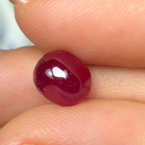 May include: A small, oval-shaped, deep red gemstone with a smooth, polished cabochon surface. The stone exhibits a rich, saturated crimson hue and a reflective, glossy finish that catches the light, highlighting its rounded, dome-like profile.