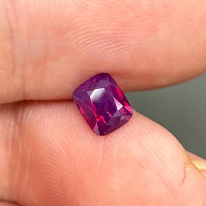 Puede incluir: Una piedra preciosa de talla cojín con un tono magenta y púrpura intenso, que presenta una superficie facetada que refleja la luz. La piedra tiene una forma rectangular con esquinas redondeadas y muestra claridad interna.