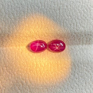 May include: Two oval-shaped, cabochon-cut star rubies with a vibrant magenta-red hue. Each gemstone displays a distinct six-rayed asterism effect, appearing as a bright white star pattern centered on the smooth, polished surface of the stones.