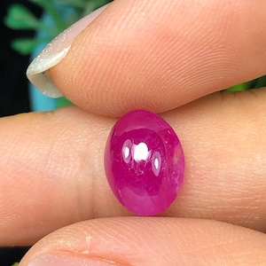 May include: A polished, oval ruby gemstone. The deep pink hue of the stone is highlighted by the light reflecting off its surface. The gemstone is held between fingers, showing its size and shape. Suitable for jewellery or collecting.