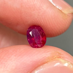 May include: A vibrant, oval-cut ruby gemstone with a deep red hue and multiple facets that reflect light. The stone displays a translucent quality with visible internal inclusions and a polished surface, showcasing a brilliant, saturated crimson color.