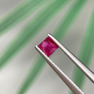 Puede incluir: Una gema roja vibrante, de corte cuadrado, es sostenida por unas pinzas plateadas sobre un fondo borroso verde y blanco. El color de la gema es rojo intenso y saturado, con texturas internas visibles. Las pinzas están enfocadas.