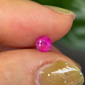 Op de afbeelding: Een ronde, gefacetteerde, felroze edelsteen. De edelsteen wordt tussen twee vingers gehouden, waarbij de briljante slijpvorm en levendige kleur worden getoond. De edelsteen lijkt een robijn te zijn, met een gepolijst oppervlak dat licht reflecteert.