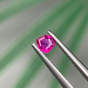 Puede incluir: Una vibrante gema rosa intenso, de corte cuadrado, es sostenida por unas pinzas plateadas. La gema tiene una superficie facetada, que refleja la luz. El fondo es verde y blanco suave, creando un contraste con el color de la gema. Esta imagen muestra una piedra preciosa.