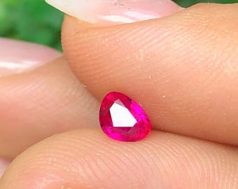 May include: A pear-shaped, faceted gemstone with a vibrant, deep pinkish-red color. The stone features a polished surface with multiple triangular and kite-shaped facets that reflect light, creating a brilliant, sparkling appearance throughout the gem.