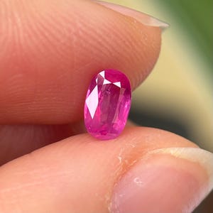 Puede incluir: Una gema ovalada de color rosa vibrante, probablemente un rubí, se sostiene entre dos dedos.