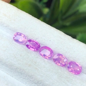 May include: Six oval-cut, light purple gemstones are arranged diagonally on a white surface. The gems have a polished, reflective surface, and the color varies slightly between each stone. The background is blurred, with green foliage visible.