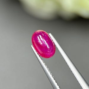 Puede incluir: Una gema vibrante de forma ovalada y corte cabujón en un tono rosa magenta intenso. La piedra presenta una superficie lisa y pulida con reflejos de luz internos y está sujeta firmemente por unas pinzas de metal plateado con puntas dentadas.