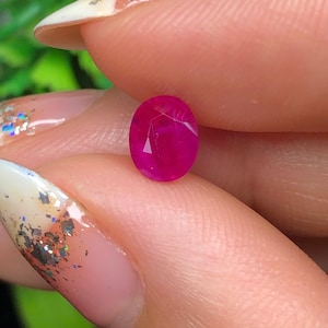 May include: A vivid, oval-shaped ruby gemstone. The gem is a deep, saturated red hue, with visible facets that reflect light. The ruby is held between fingers, highlighting its polished surface and clarity. The background is blurred, focusing on the gemstone.