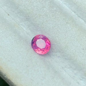 May include: A round, faceted gemstone with a vibrant pink and magenta hue. The stone features a brilliant cut with multiple triangular and kite-shaped facets that reflect light, creating a sparkling appearance across its polished surface.