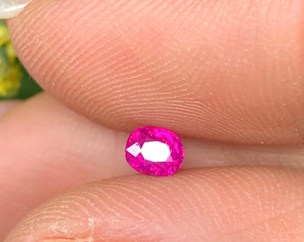 May include: A vibrant, oval-cut, hot pink gemstone. The gem is held between two fingers, showcasing its polished facets and internal brilliance. The stone's color is a deep, saturated pink, reflecting light. This gemstone is likely for jewelry making.