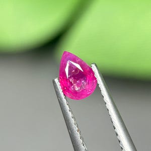 Op de afbeelding: Een levendige, peervormige losse edelsteen met een diepe fuchsia roze tint en een gefacetteerd oppervlak. De steen wordt vastgehouden door een zilverkleurige metalen pincet, wat de doorschijnende kwaliteit en de scherpe punt duidelijk toont.